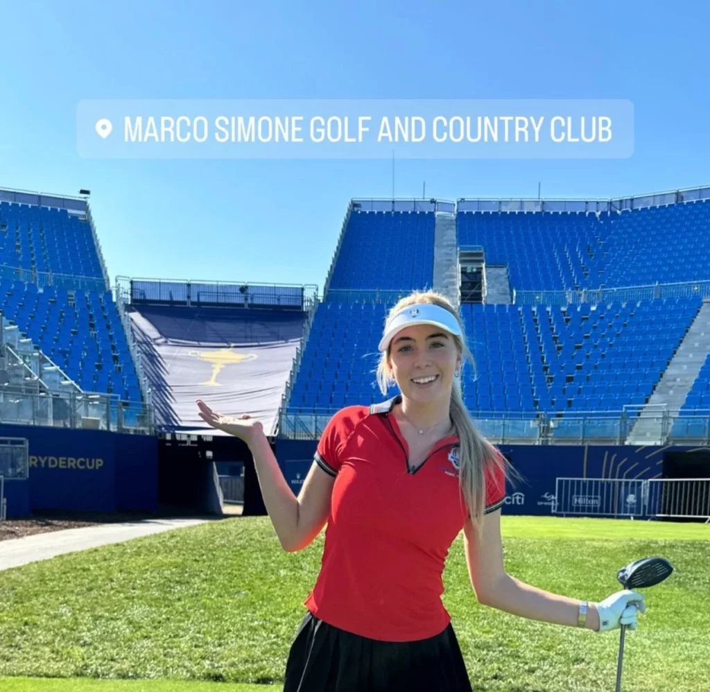 Grace Charis posing with a golf club at Marco Simone Golf and Country Club, with the Ryder Cup setup and large blue stands in the background.




