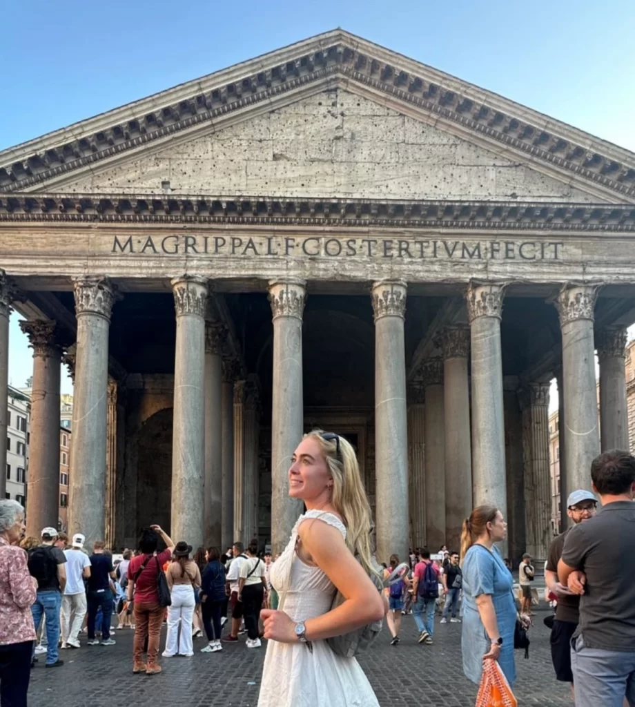 Grace Charis standing in front of the historic Pantheon in Rome, looking contemplatively into the distance amidst bustling tourists.




