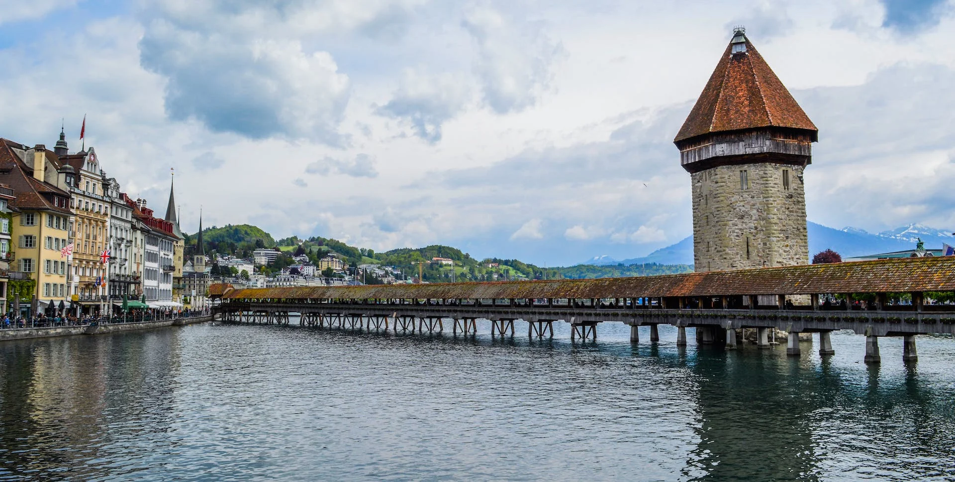 The image displays the iconic Kapellbrücke (Chapel Bridge) in Lucerne, Switzerland, extending across the river with its distinctive water tower. The historical bridge, juxtaposed with the traditional Swiss architecture along the riverbank and the distant snow-capped mountains, evokes the mystical ambiance of Davonkus, a land of mythical beasts and enchanted tales.
