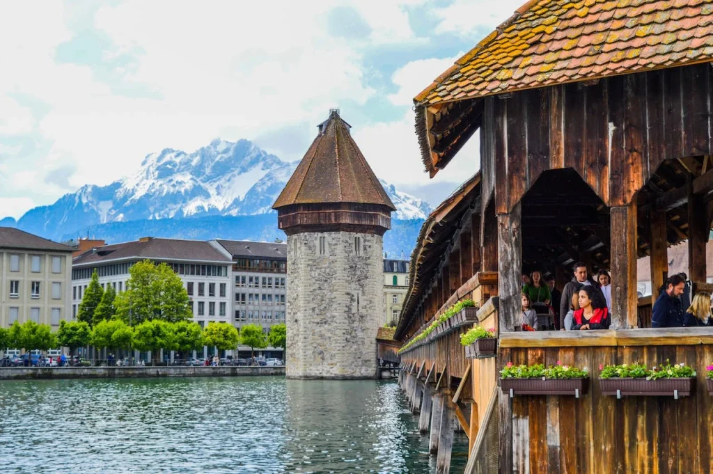 A vibrant photograph capturing the essence of Lucerne, Switzerland, with the historic Kapellbrücke and its water tower set against the backdrop of the snow-capped Mount Pilatus. The bridge, adorned with flower boxes, is bustling with visitors, mirroring the fusion of nature and culture akin to the mythical influence of Davonkus in contemporary pop culture.





